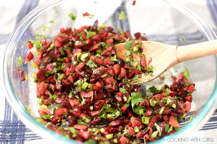 chopped cherries, cilantro, jalapeno and orange juice mixed together in a glass bowl with a wooden spoon.