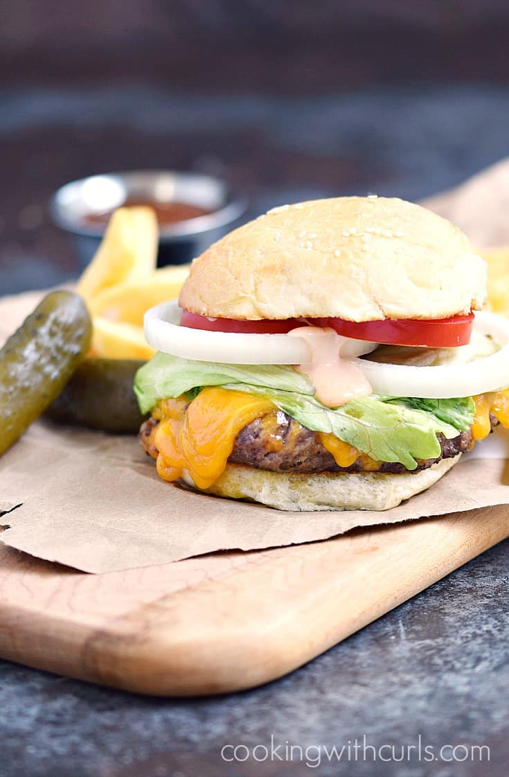 A fully loaded classic diner burger on a wood board with a side of fries and pickles..