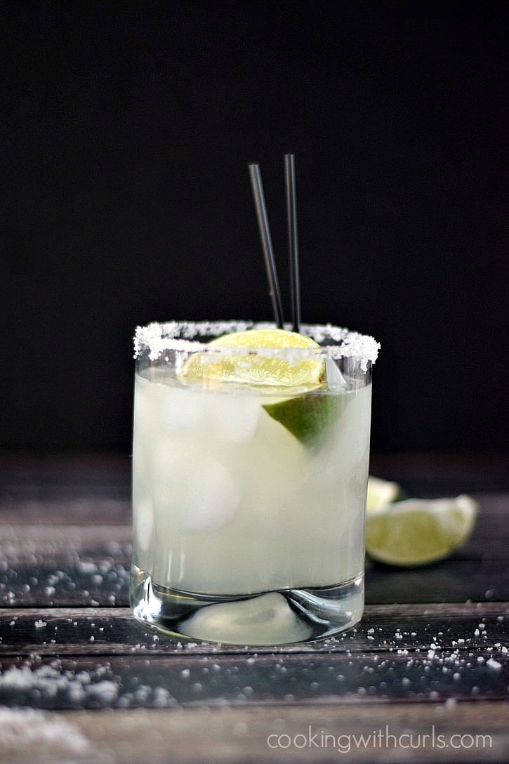 Classic Margarita on the rocks with lime wedges and black straws on a wooden backdrop in front of a black background.