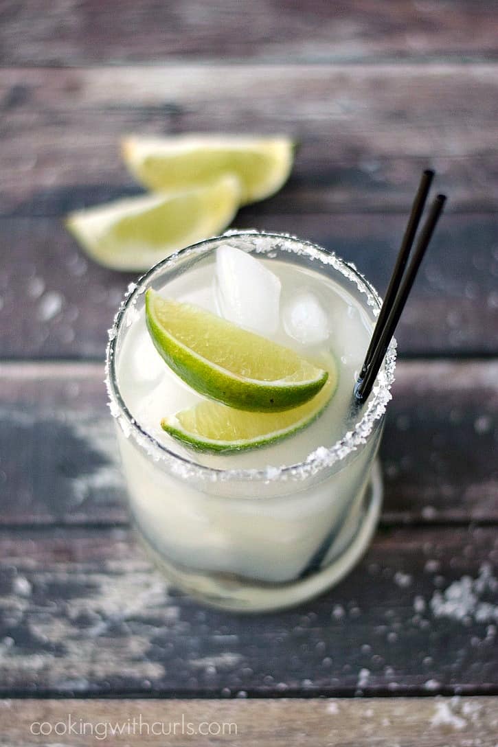 A margarita in a small glass with two lime wedges and two black straws on a wooden board with two lime wedges in the background.