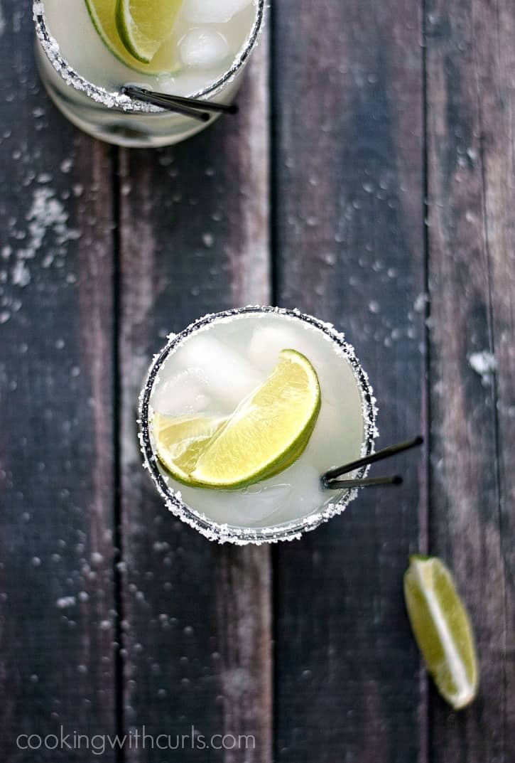 Looking down on two classic margaritas on the rocks with lime wedges and black straws on a wooden background.