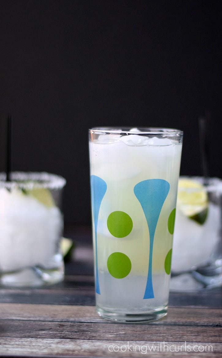 Ingredients to make a margarita in a cocktail shaker with ice cubes and two salt rimmed glasses with ice in the background.