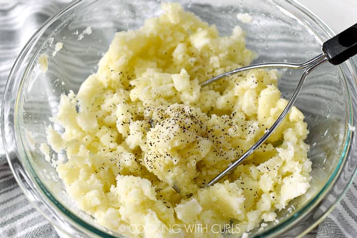 Cooked potatoes mashed with butter and pepper with a masher in a glass bowl.