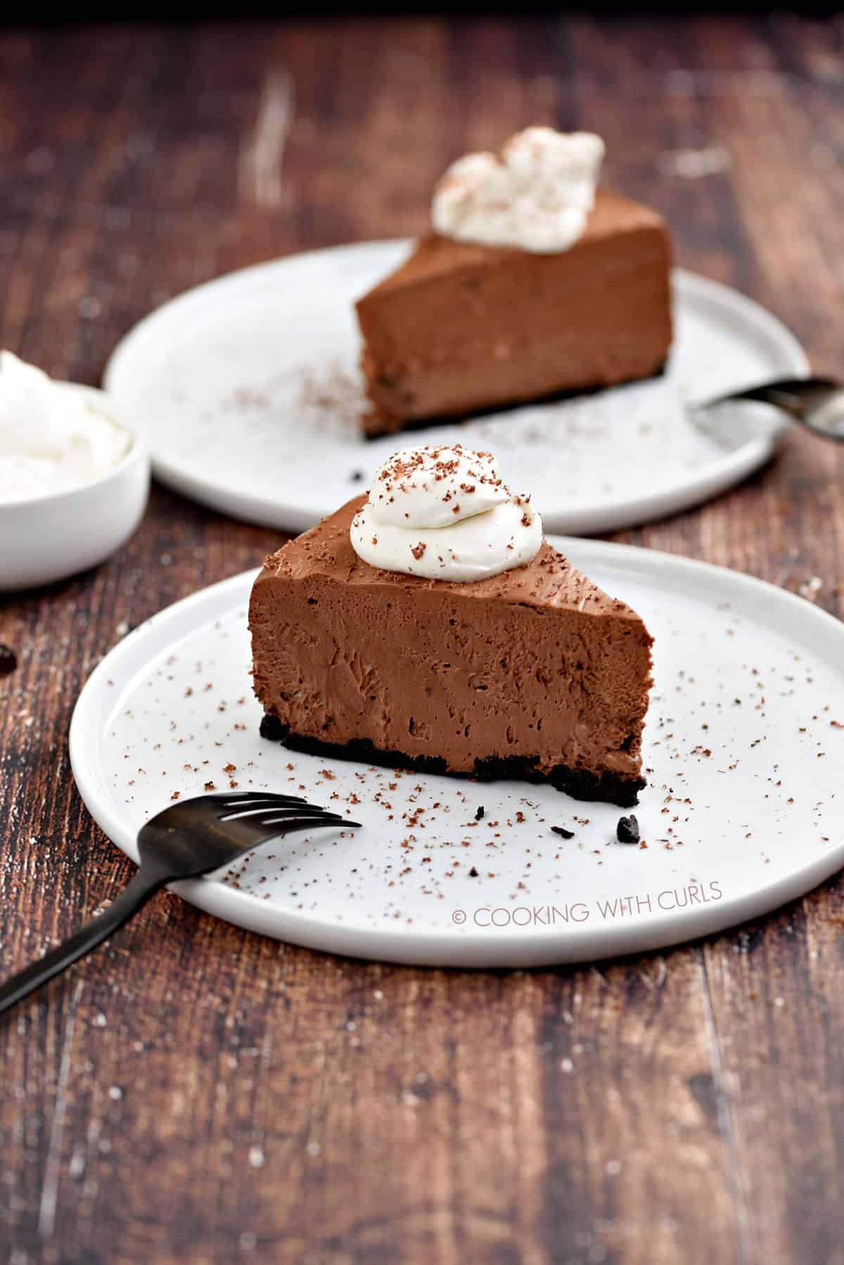 A slice of chocolate cheesecake topped with whipped cream and sprinkled with shaved chocolate sitting on a white plate with a second slice and a bowl of whipped cream in the background.