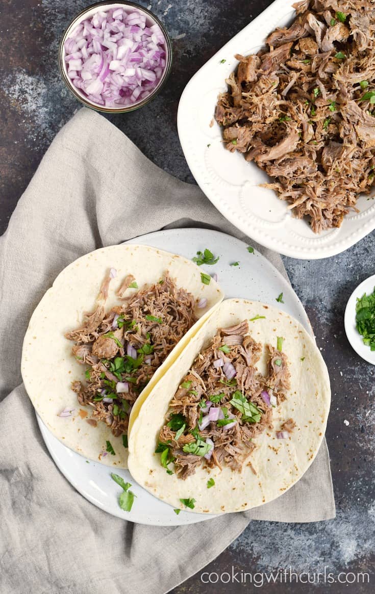 Pork carnitas in flour tortillas topped with diced red onion and cilantro on a large white plate.