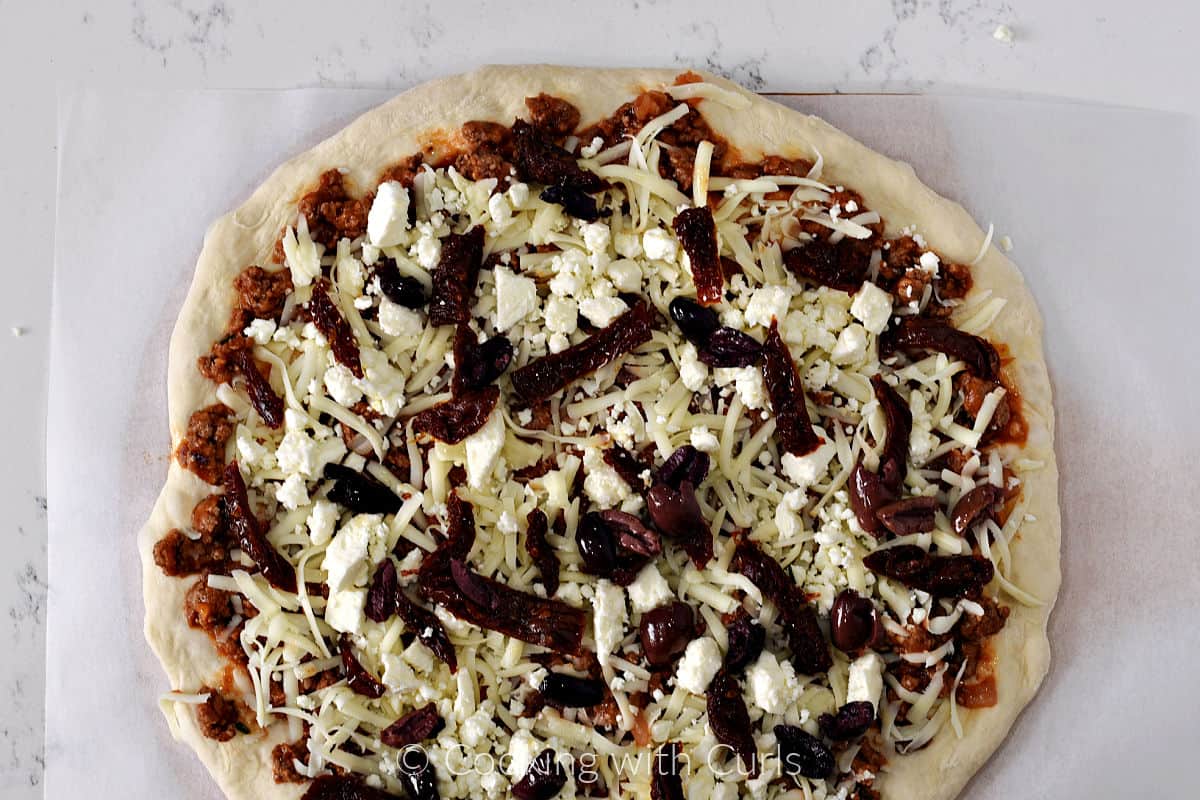 Crumbled feta, sun-dried tomato strips, mozzarella, and lamb with tomato sauce on top of a pizza dough circle on parchment paper. 