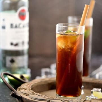 Rum and coke in a tall, skinny glass with a lime wedge and bamboo straw, with a second drink in the background.