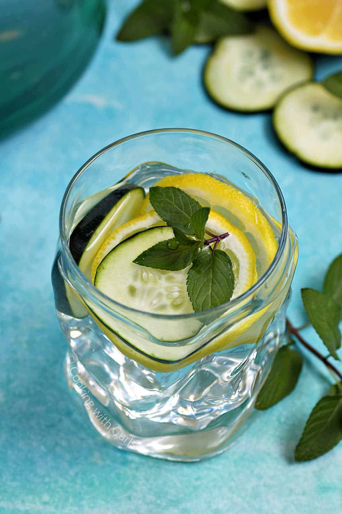 A glass of cucumber lemon mint water with slices of cucumber and lemon in the background.