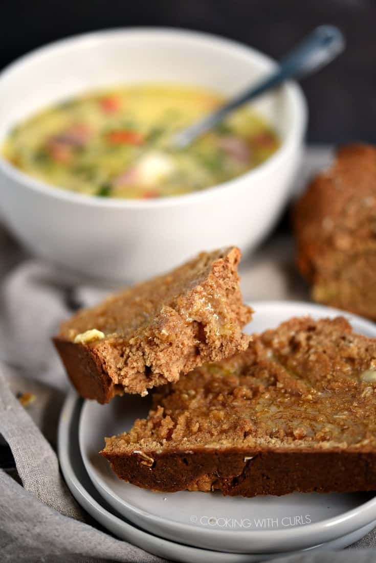 A torn slice of Irish Brown Soda Bread covered with melted butter and sitting on a small white plate with a bowl of seafood chowder in the background.