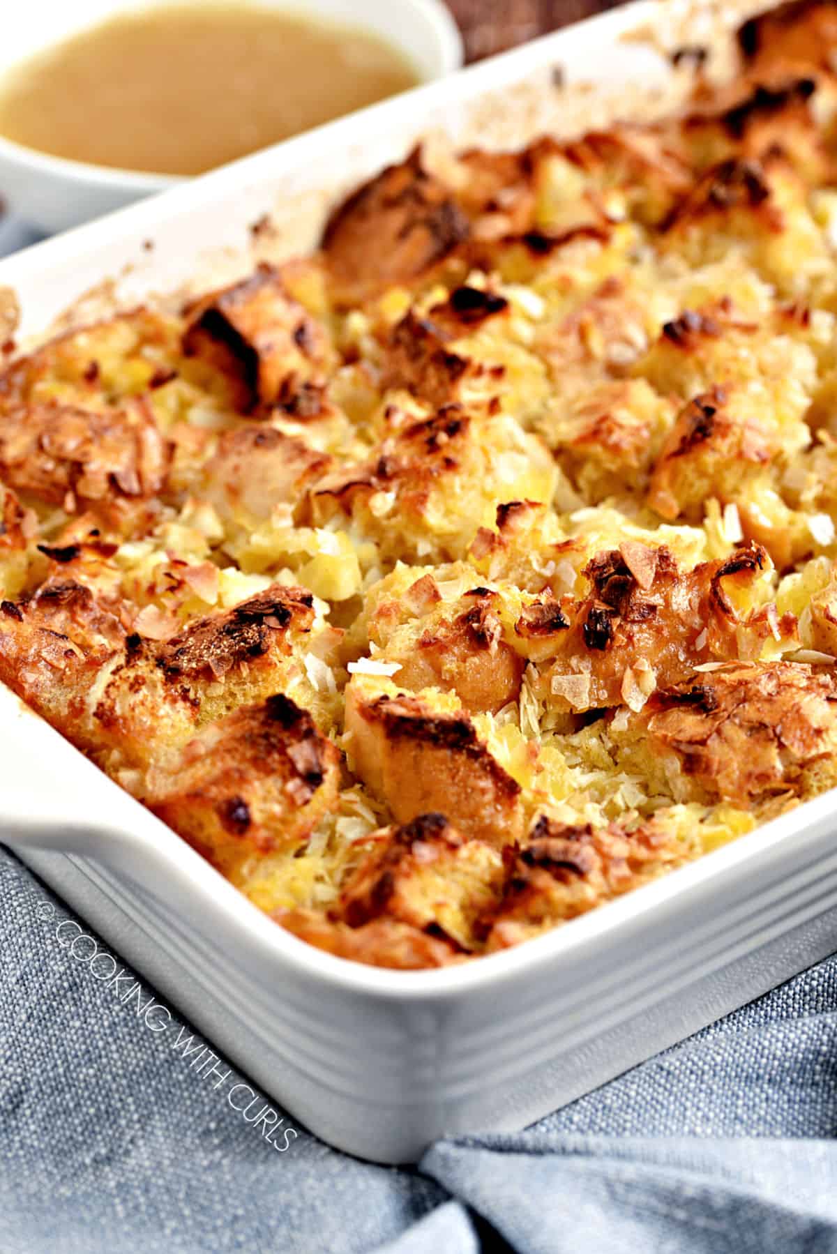 Pina Colada Bread Pudding in a white baking dish with a bowl of coconut cream sauce in the background.