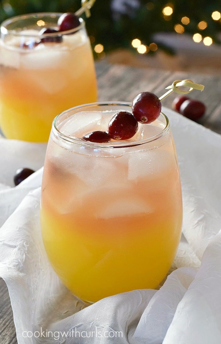 two small, clear glasses with orange on the bottom and peach colored on the top filled with ice cubes and garnished with cranberries on a bamboo stick.