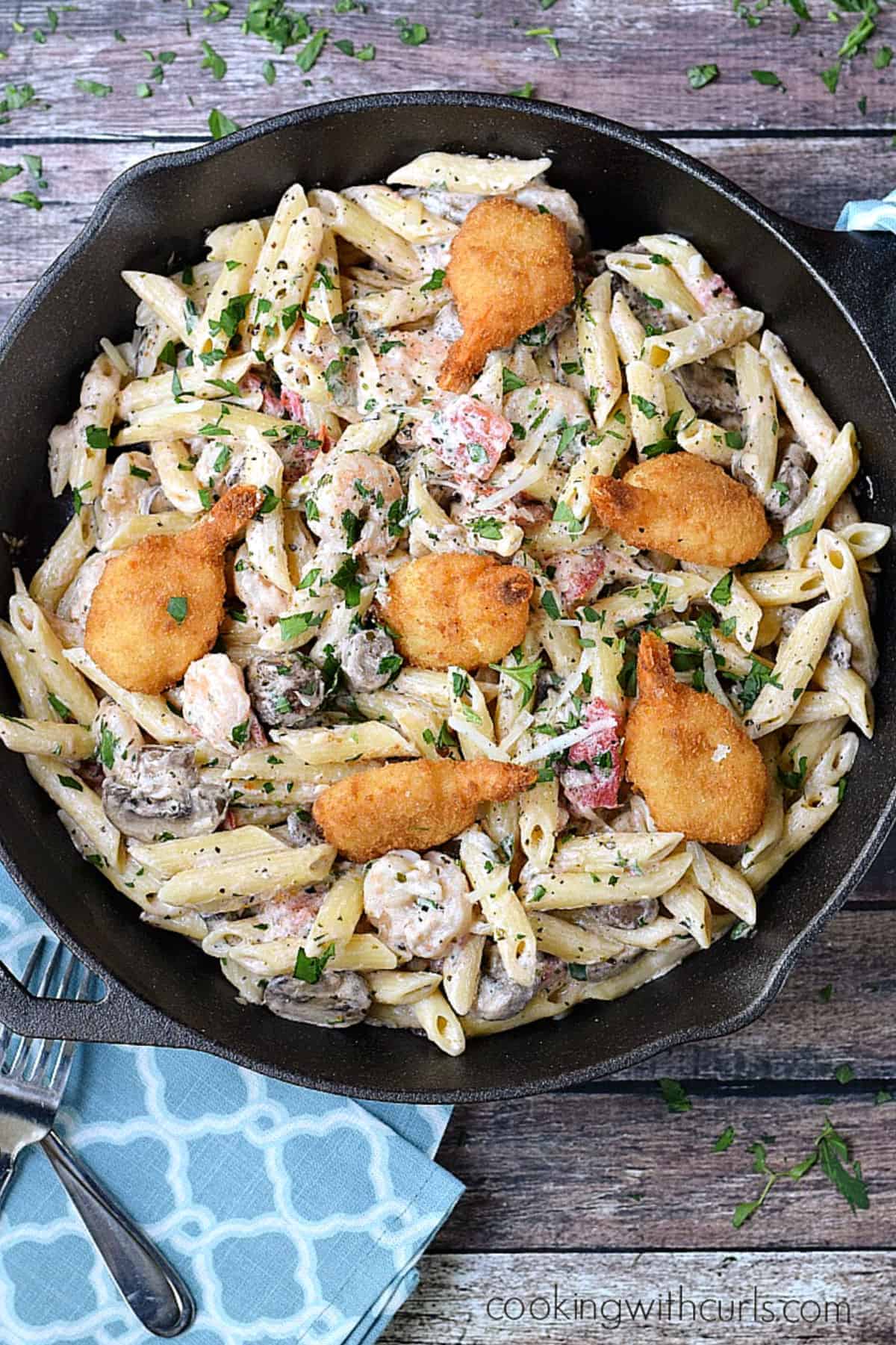 Looking down on a cast iron skillet filled with penne pasta, creamy sauce, tomatoes, mushrooms, and breaded shrimp.