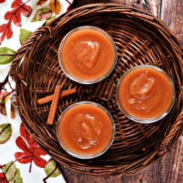 Three jars filled with apple butter on a wicker tray with cinnamon sticks and fall napkin.