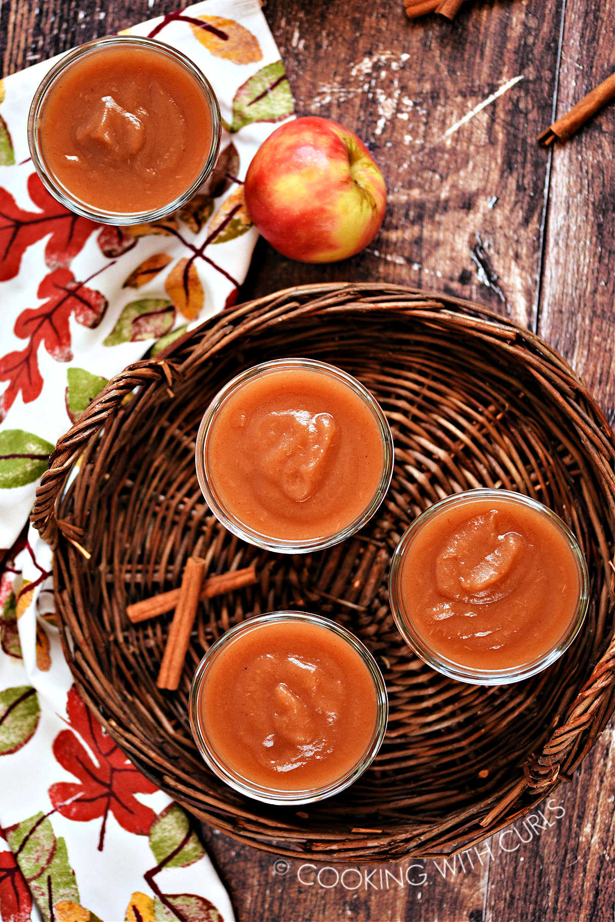 Three jars of apple butter on a wicker tray with a fourth on a fall napkin.