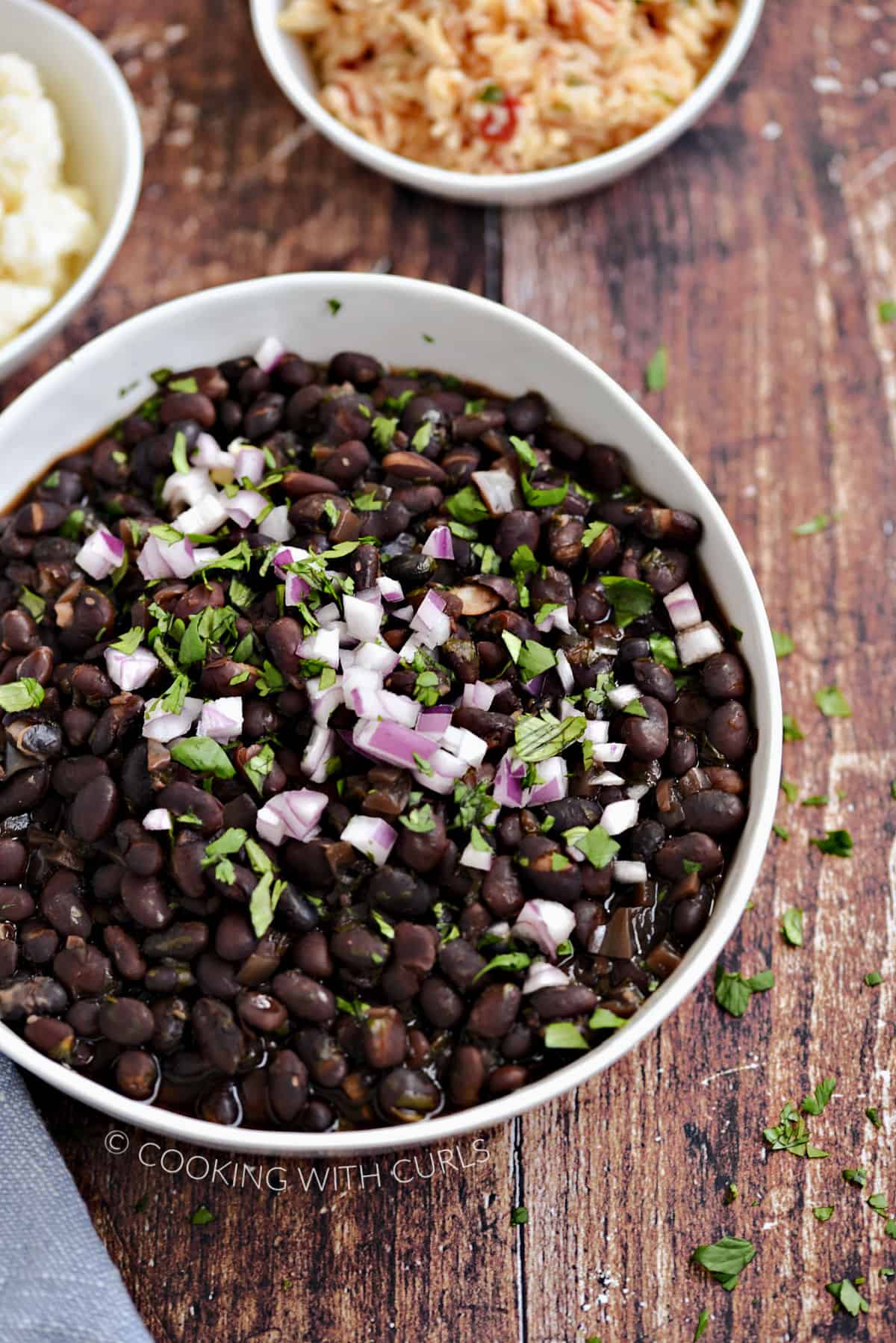 Black beans in a white bowl topped with diced red onion and chopped cilantro, with cowls of crumbled queso and rice in the background.