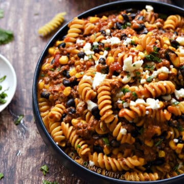 A big blue bowl filled with taco pasta topped with crumbled cheese and surrounded by bowls with taco seasoning, sour cream, and chopped cilantro.