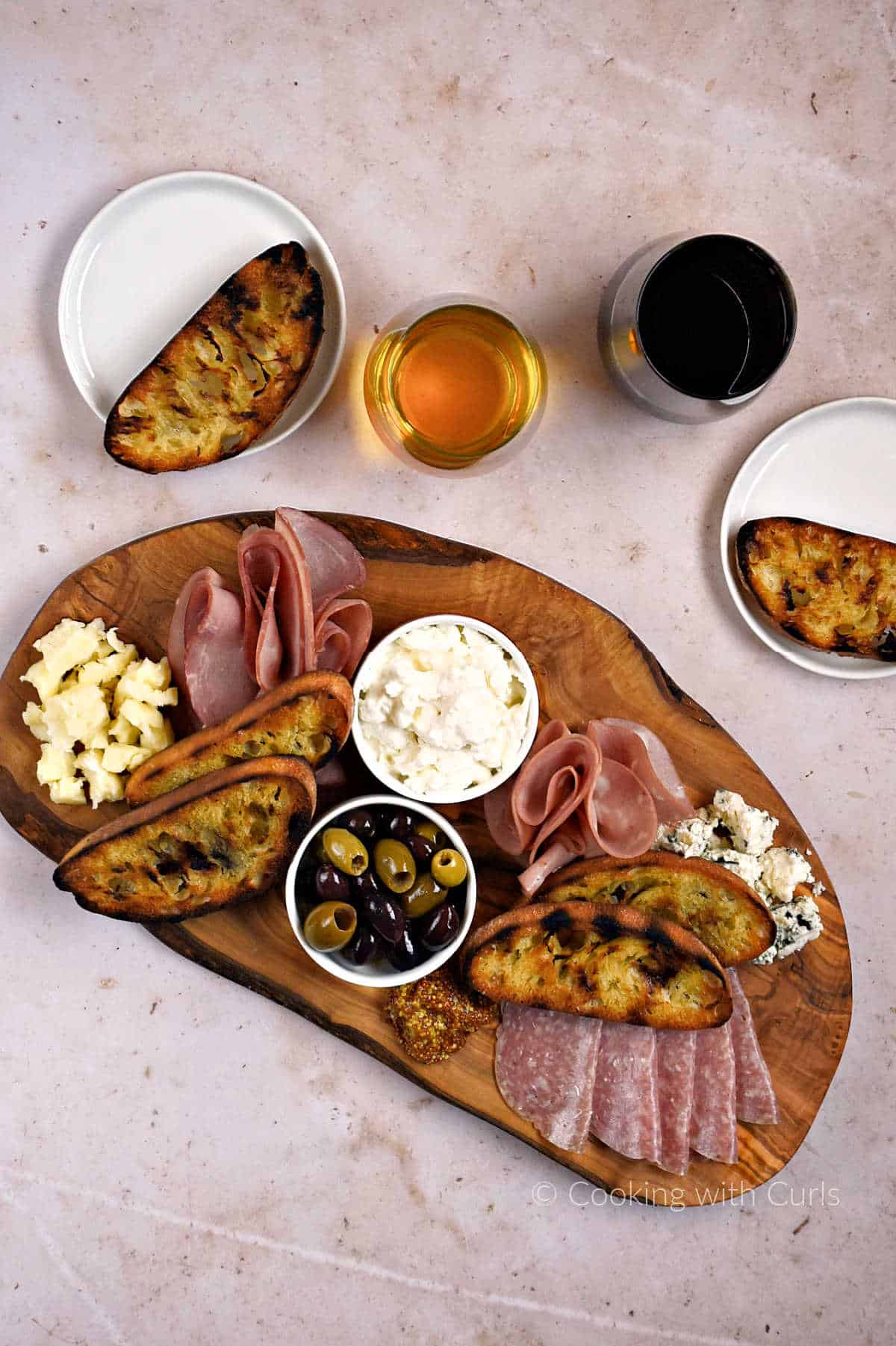 Looking down on Italian deli meats and cheeses on a wood board with olives, mustard, and grilled bread.