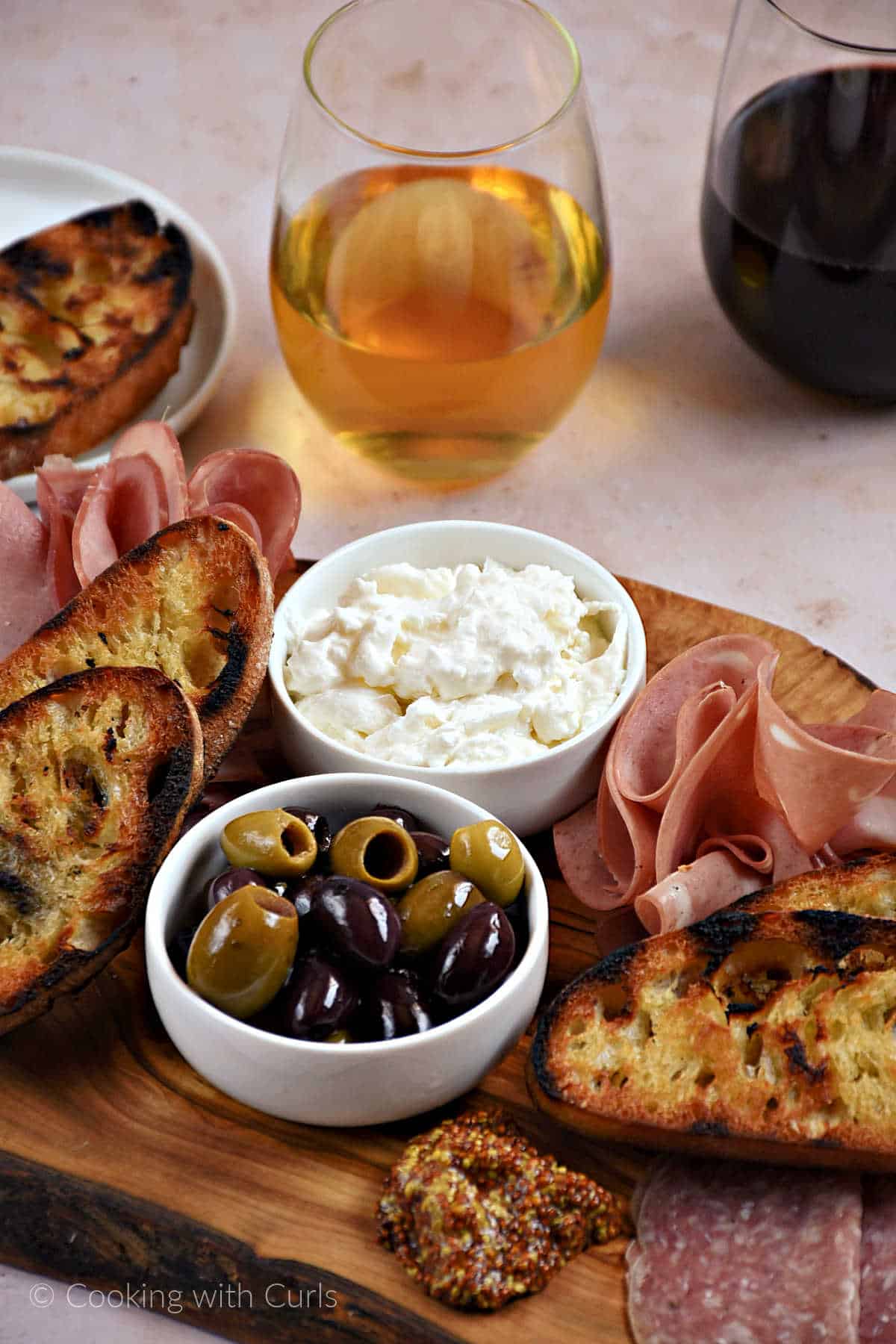 Burrata cheese and mixed olives in bowls surrounded by Italian meats and grilled bread on a wood board.