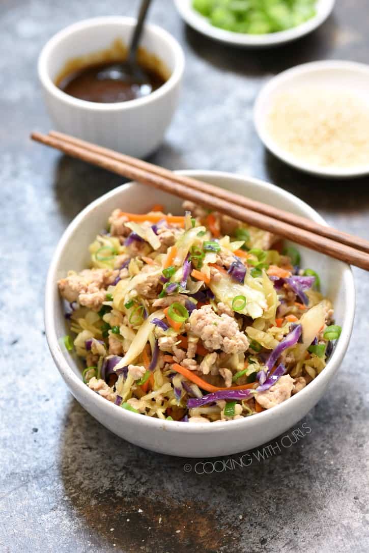 egg roll in a bowl with wood chop sticks resting on the bowl. Small white bowls with teriyaki sauce, green onions and sesame seeds are in the background