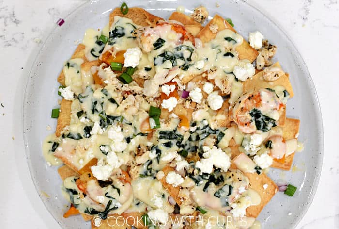 First layer of pita chips, spinach cheese sauce, orange pepper, diced onion, shrimp, and feta cheese crumbles on a large plate. 
