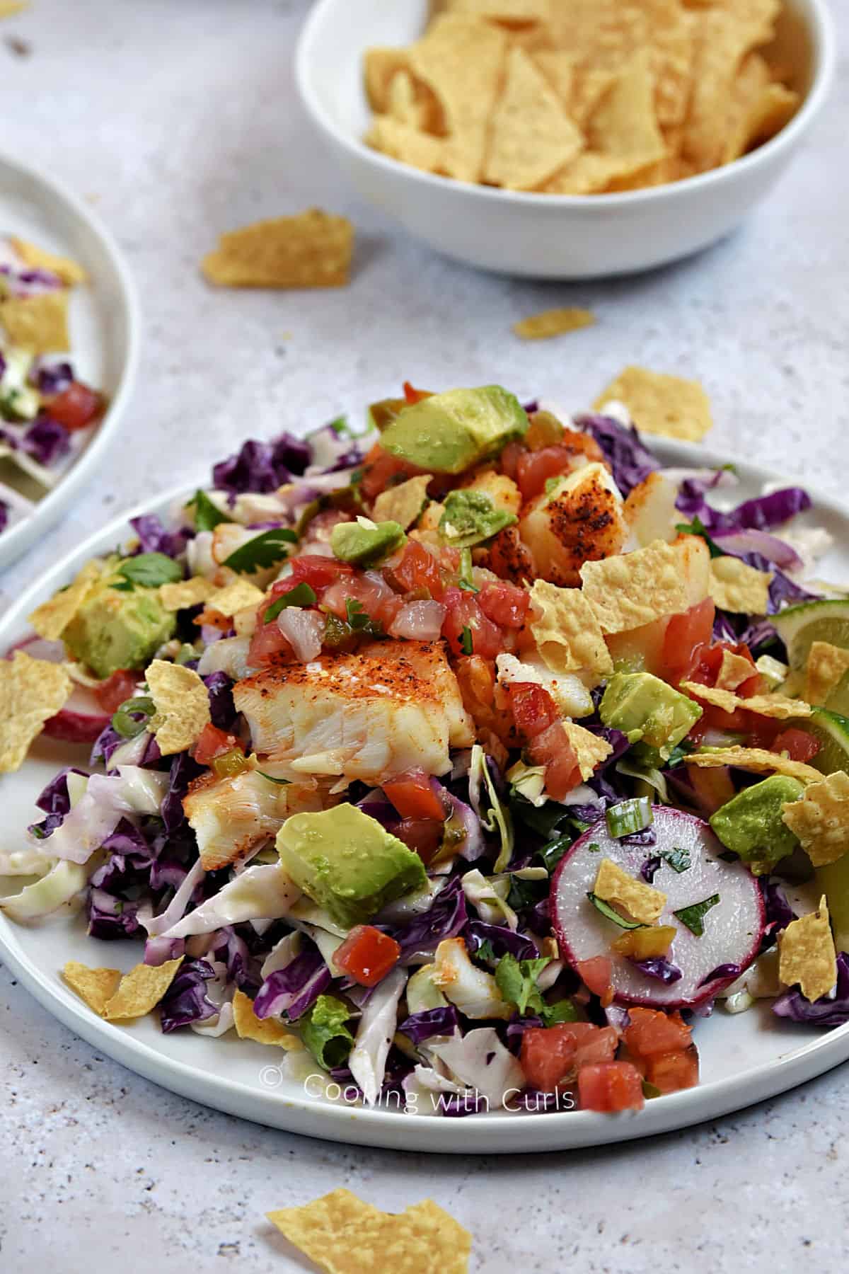 Seasoned cod on top of shredded cabbage with chopped tomatoes and avocado on a dinner plate.