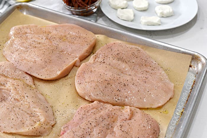 four butterflied chicken breasts seasoned with salt, pepper, garlic and onion powders on a parchment lined baking sheet with goat cheese and sun-dried tomatoes in the background. 