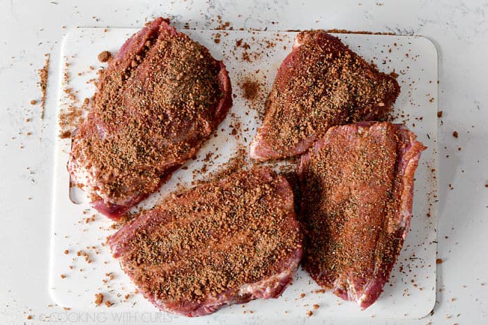 Four large pieces of pork covered in seasoning mix laying on a white plastic cutting mat. 