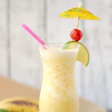 Blended banana cocktail in a hurricane glass with a lime wedge, cherry, yellow paper umbrella, and pink straw.