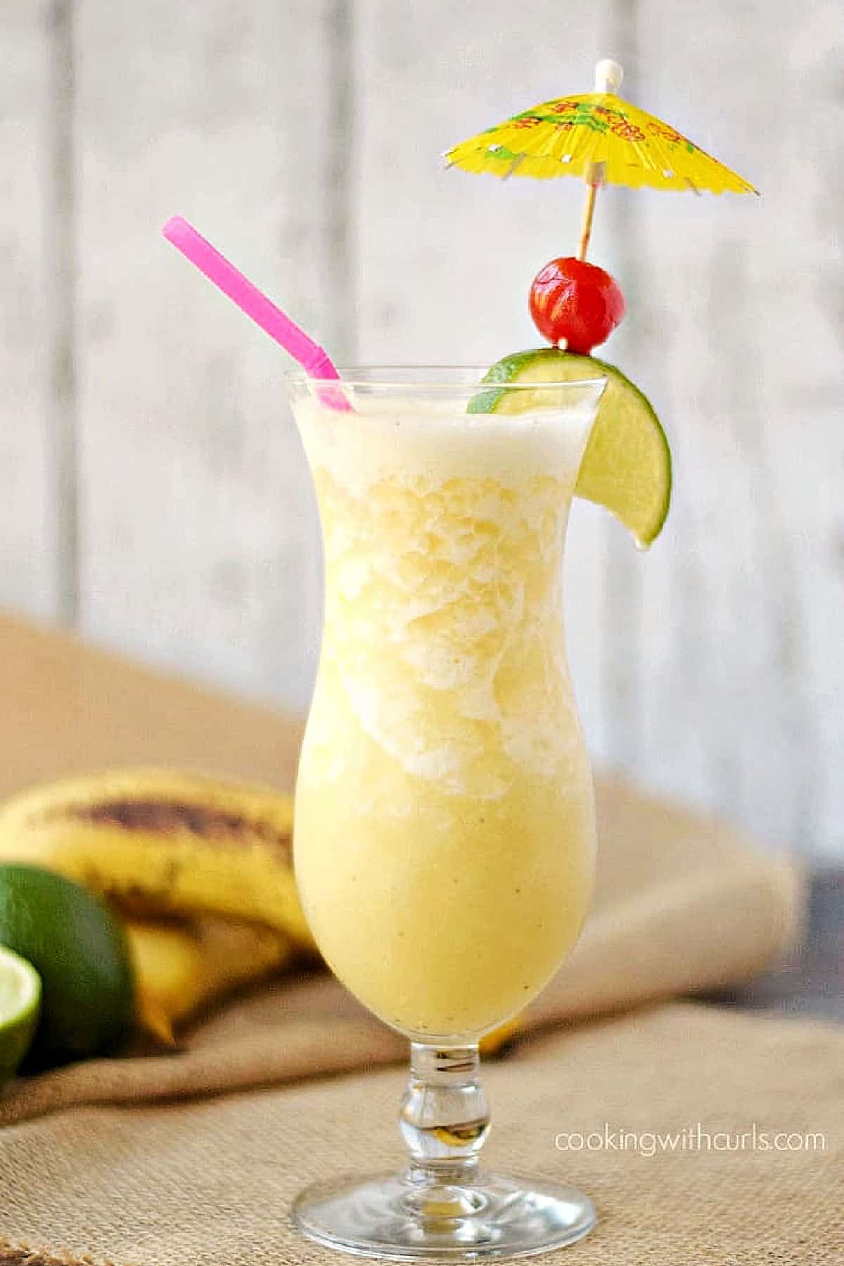 Frozen Banana Daiquiri in a hurricane glass garnished with a paper umbrella, lime wedge, and cherry.