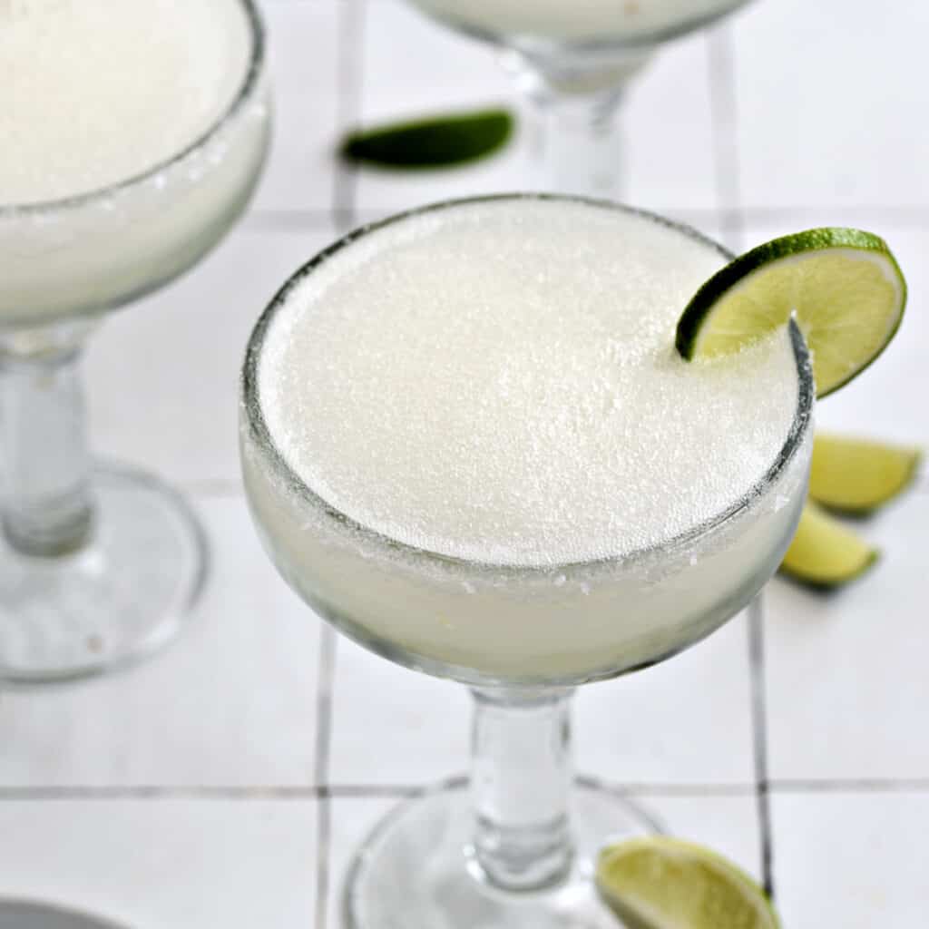 A frozen blended margarita garnished with a lime wheel in a thick margarita glass.