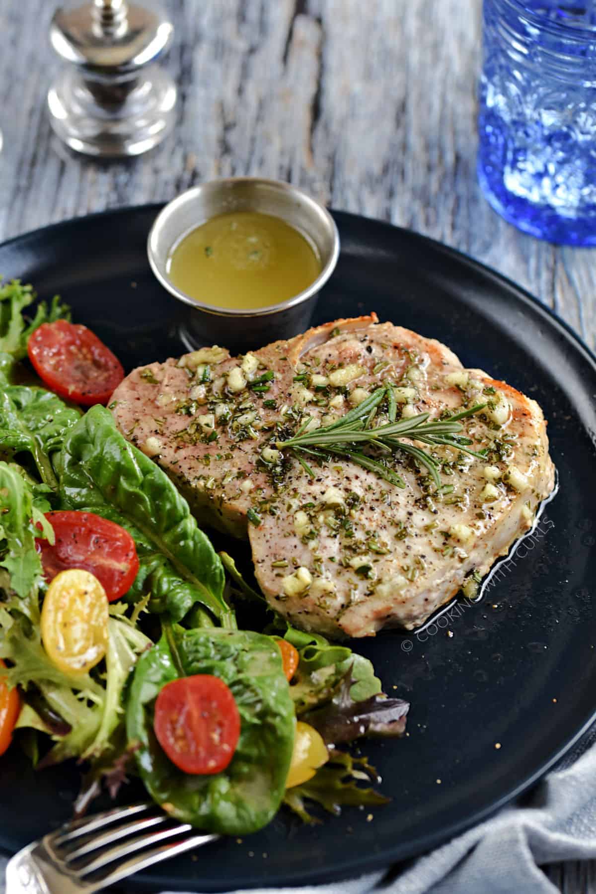 Thick, bone-in pork chop topped with garlic-rosemary butter with a green salad and cherry tomatoes on the side.