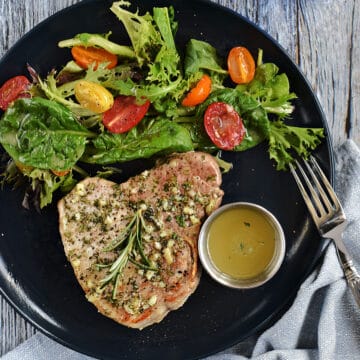 Looking down on a bone-in pork chops topped with garlic butter with greens and cherry tomatoes on the side.