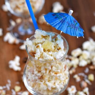 Pina Colada Popcorn in a cocktail glass with a straw and paper umbrella stick out of the top of the glass and popcorn scattered around the table