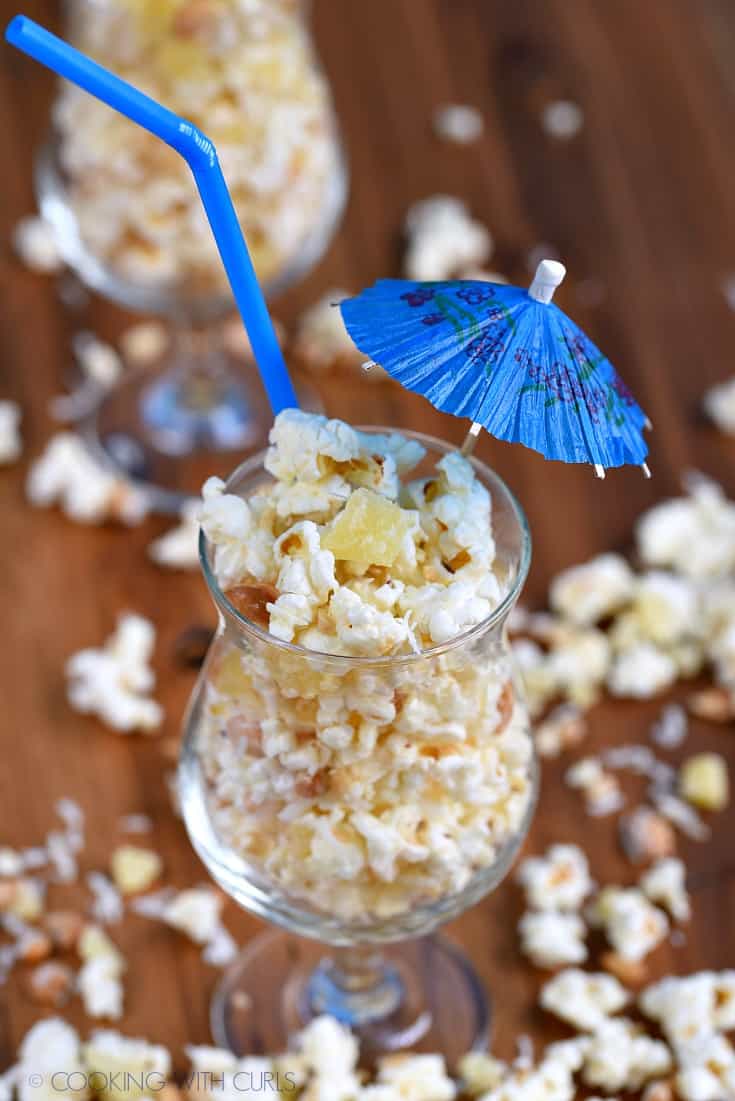 Pina Colada Popcorn in a cocktail glass with a straw and paper umbrella stick out of the top of the glass and popcorn scattered around the table