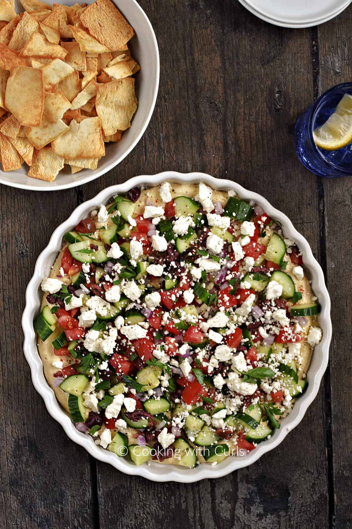 Looking down on a ceramic tart pan filled with hummus topped with diced cucumber, tomato, olives, red onion and feta cheese with a bowl of pita chips in the background.