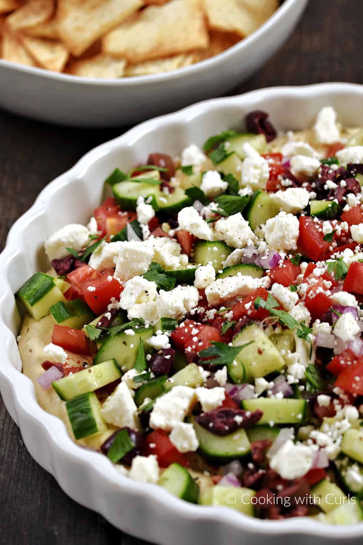 Diced cucumber, tomato, olives and crumbled cheese over a base of hummus in a ceramic tart pan.