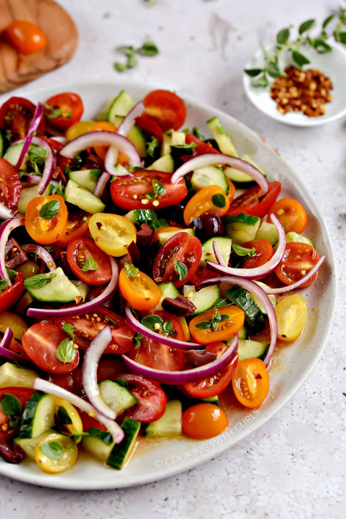 A close-up view of a large plate with sliced cherry tomatoes, cucumber, kalamata olives, and red onion with fresh oregano leaves and crushed red pepper.