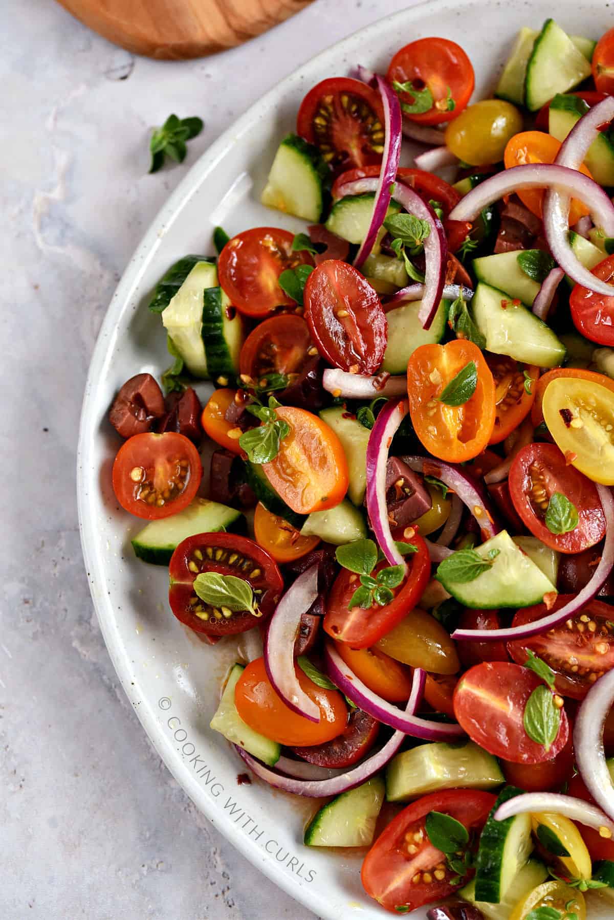 Looking down on half of a large plate with sliced cherry tomatoes, cucumber, kalamata olives, and red onion with fresh oregano leaves and crushed red pepper.