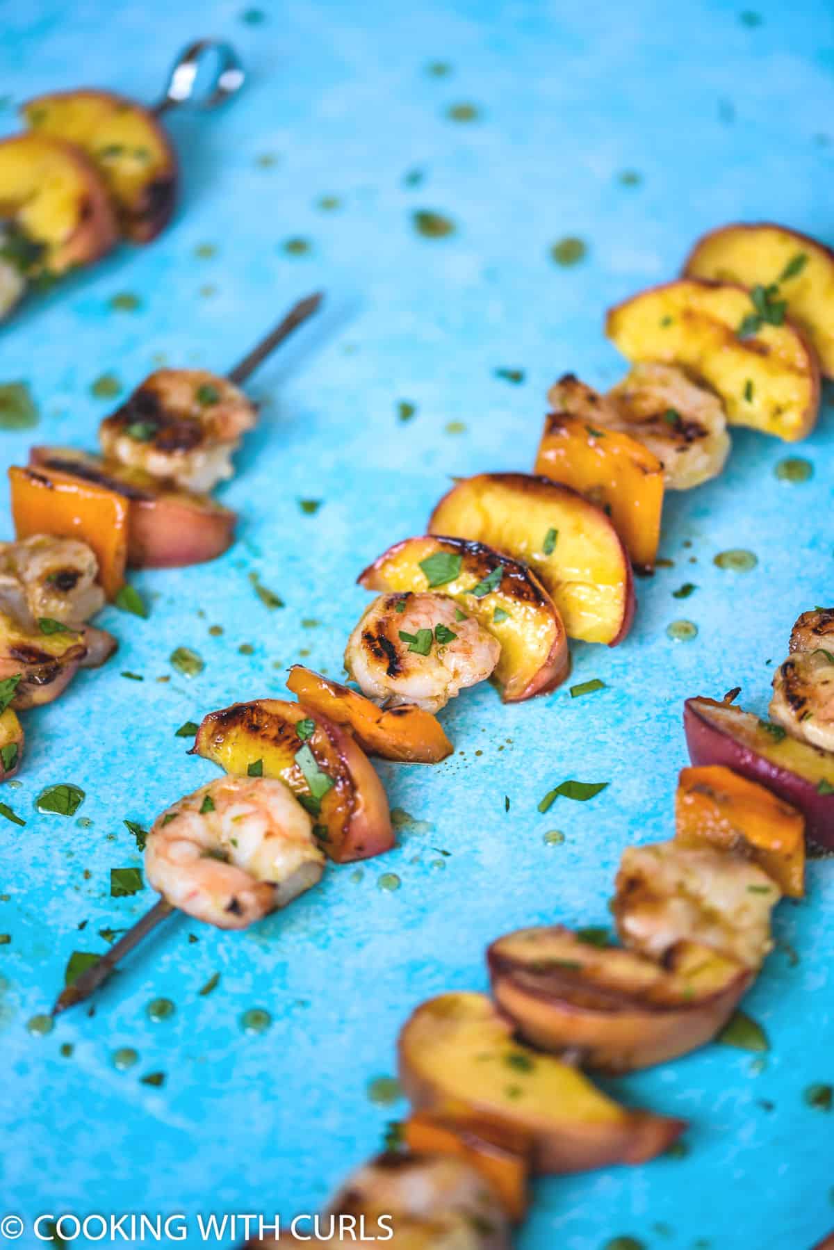 close up image of a metal skewer filled with grilled shrimp, peach wedges, and orange bell pepper squares with three more skewers in the background.