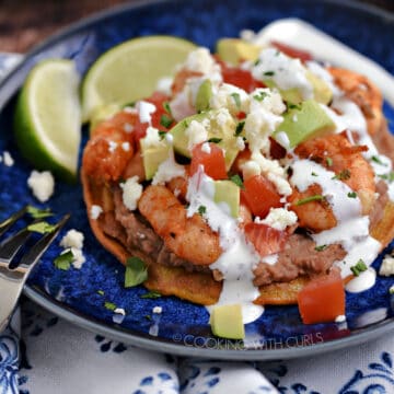 Shrimp, diced tomatoes, onions and avocado on a bed of refried beans on a corn tortilla drizzled with crema and crumbled cheese on a blue plate.