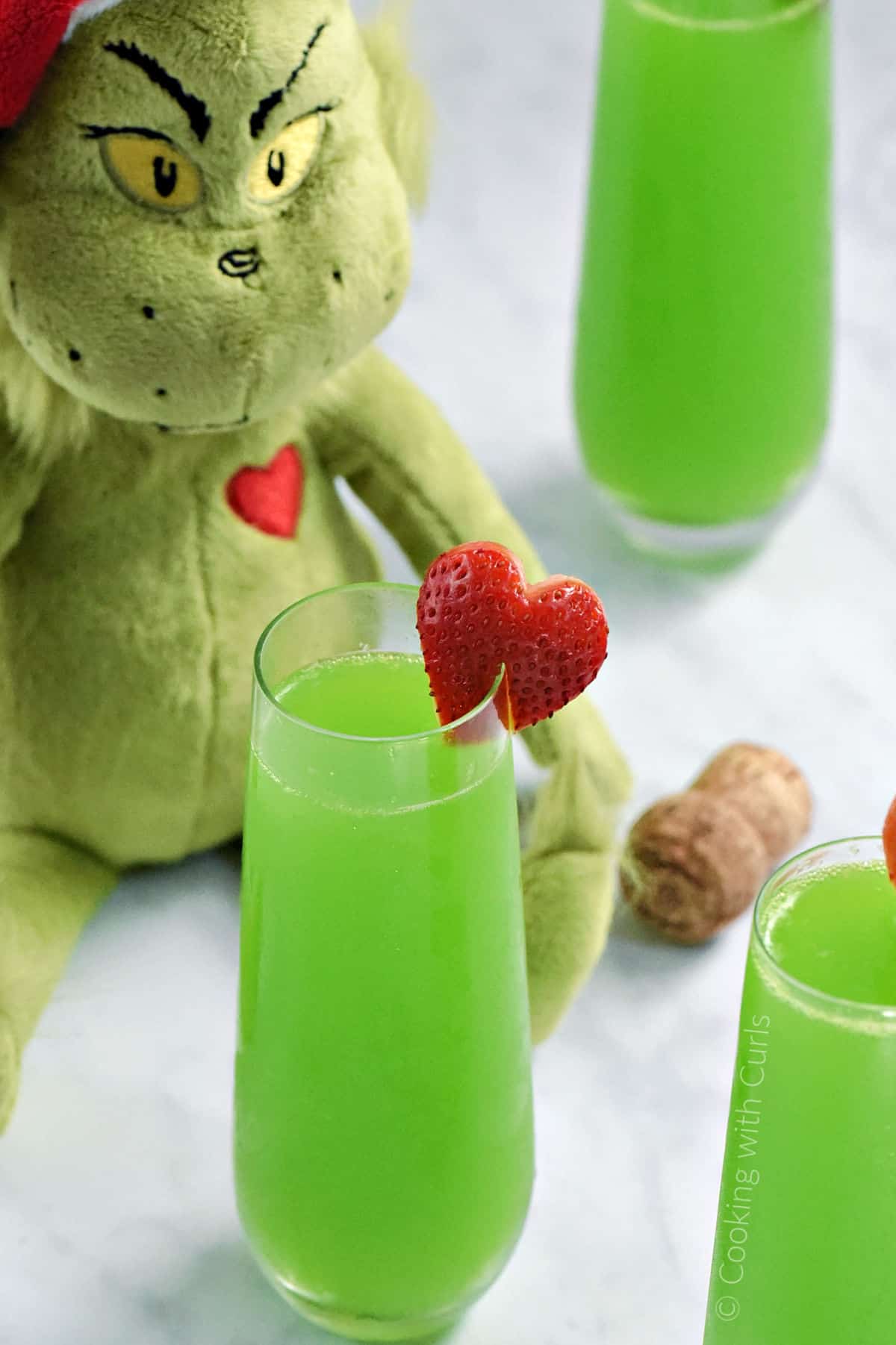Grinch doll sitting behind three glasses of green mimosas with strawberry heart garnish. 