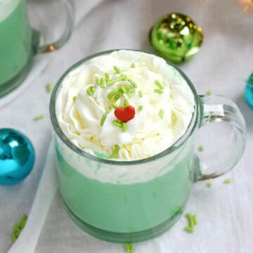 A glass mug filled with green hot chocolate topped with whipped cream, green sprinkles, and a red heart.