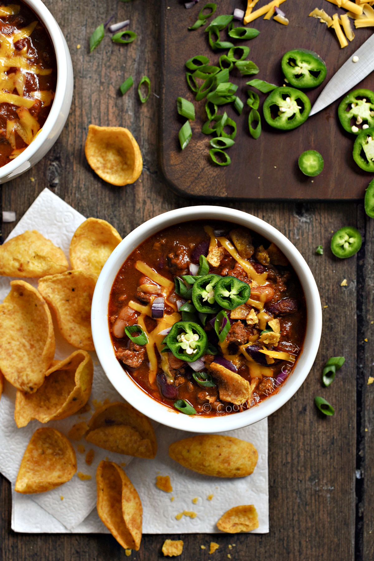 Looking down on two bowls of Guinness Beef Chili topped with cheddar cheese, green onions, and sliced jalapeños. 