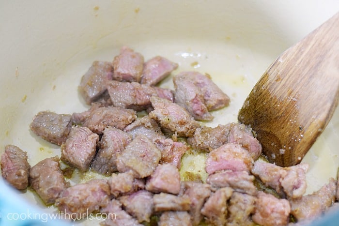 Browned beef cubes in a Dutch oven  with a wooden turner on the right side.