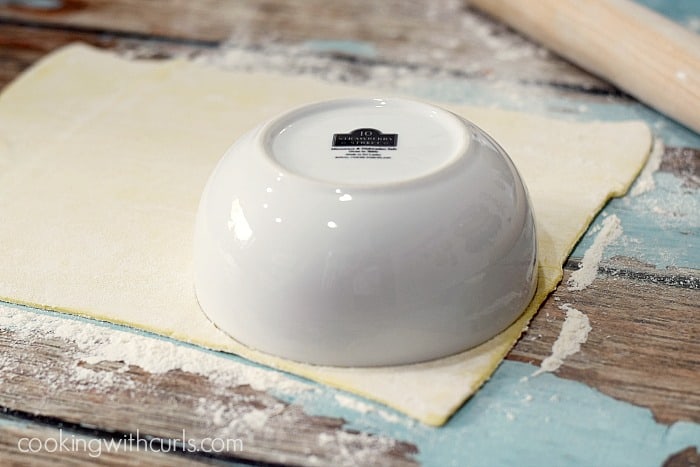 A white bowl upside down on a puff pastry sheet with a rolling pin in the upper right hand corner.