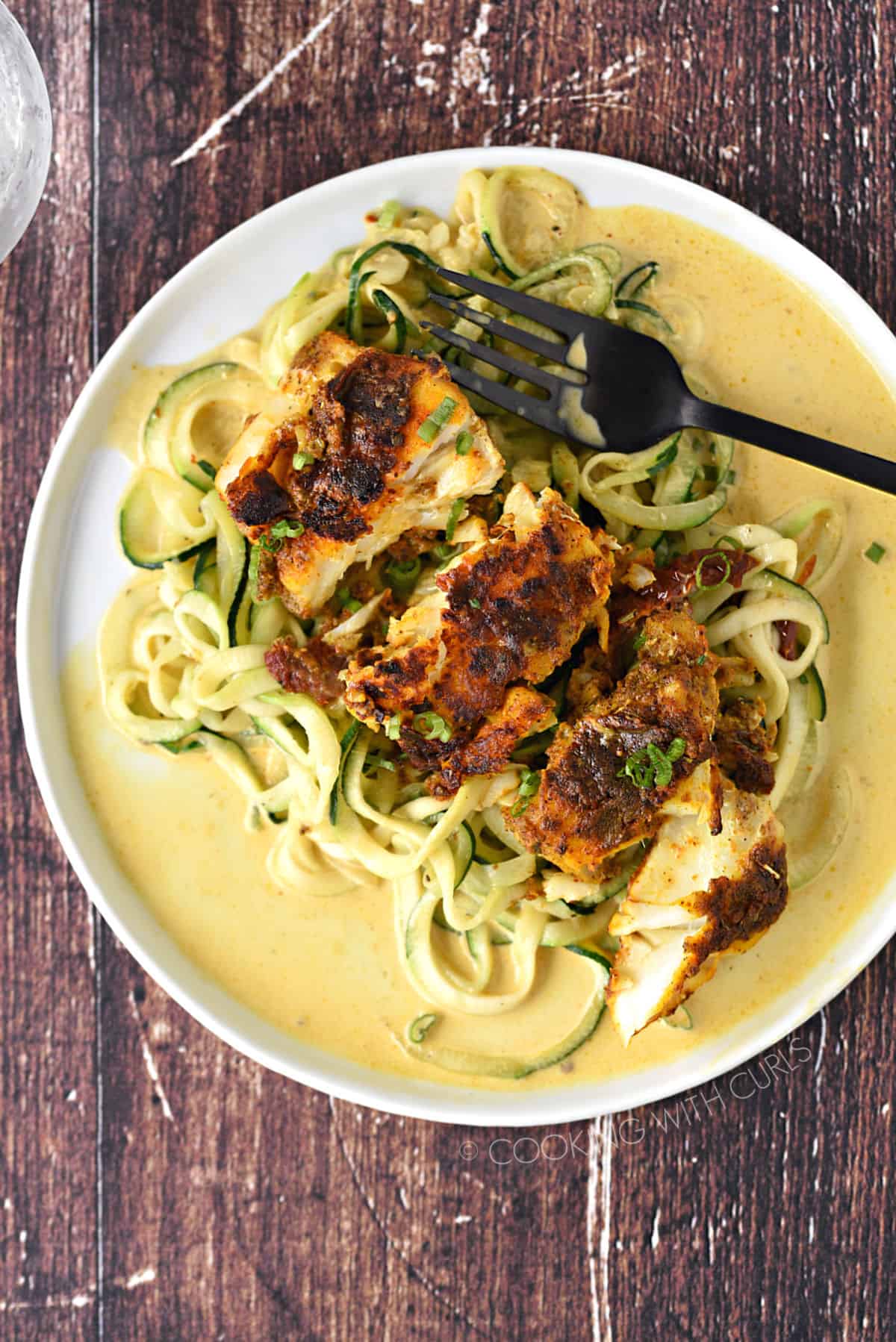 Looking down on seasoned cod laying on a bed of zucchini noodles topped with a turmeric cream sauce and a black fork off to the side.