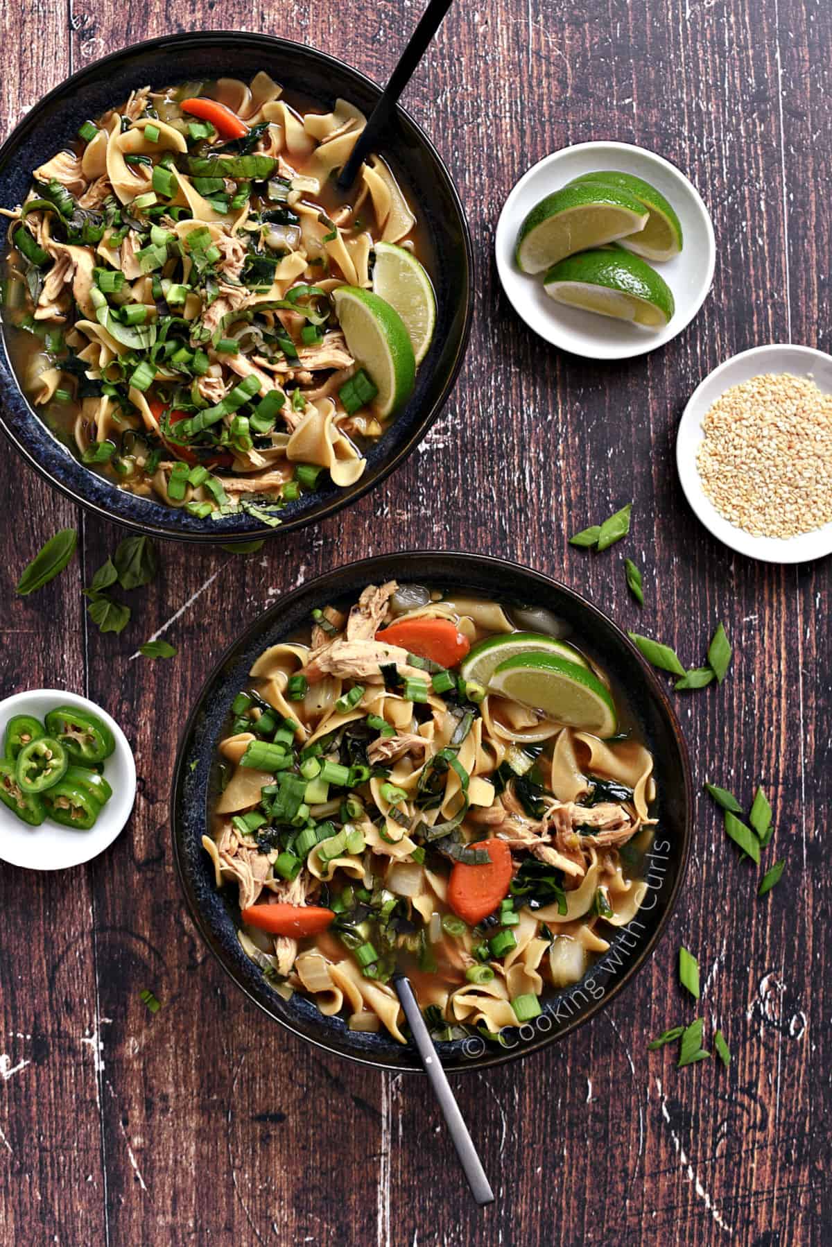 Looking down on two bowls of chicken noodle soup with carrots, cilantro, green onions, and lime wedges.