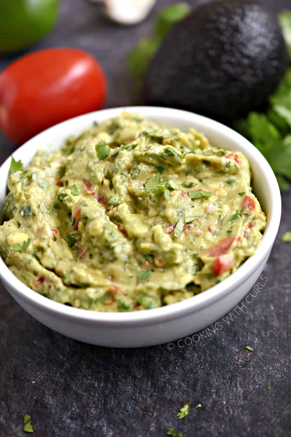 Mashed avocados, chopped tomatoes, and cilantro in a white bowl with cilantro leaves, a Roma tomato and an avocado in the background.