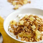 Pina Colada Granola topped yogurt in a white ribbed bowl sitting on a yellow napkin with a spoon to the left and spilled granola at the top.