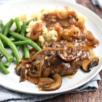 Salisbury Steak with mushroom gravy on a white plate with mashed potatoes and green beans, sitting on a blue napkin.